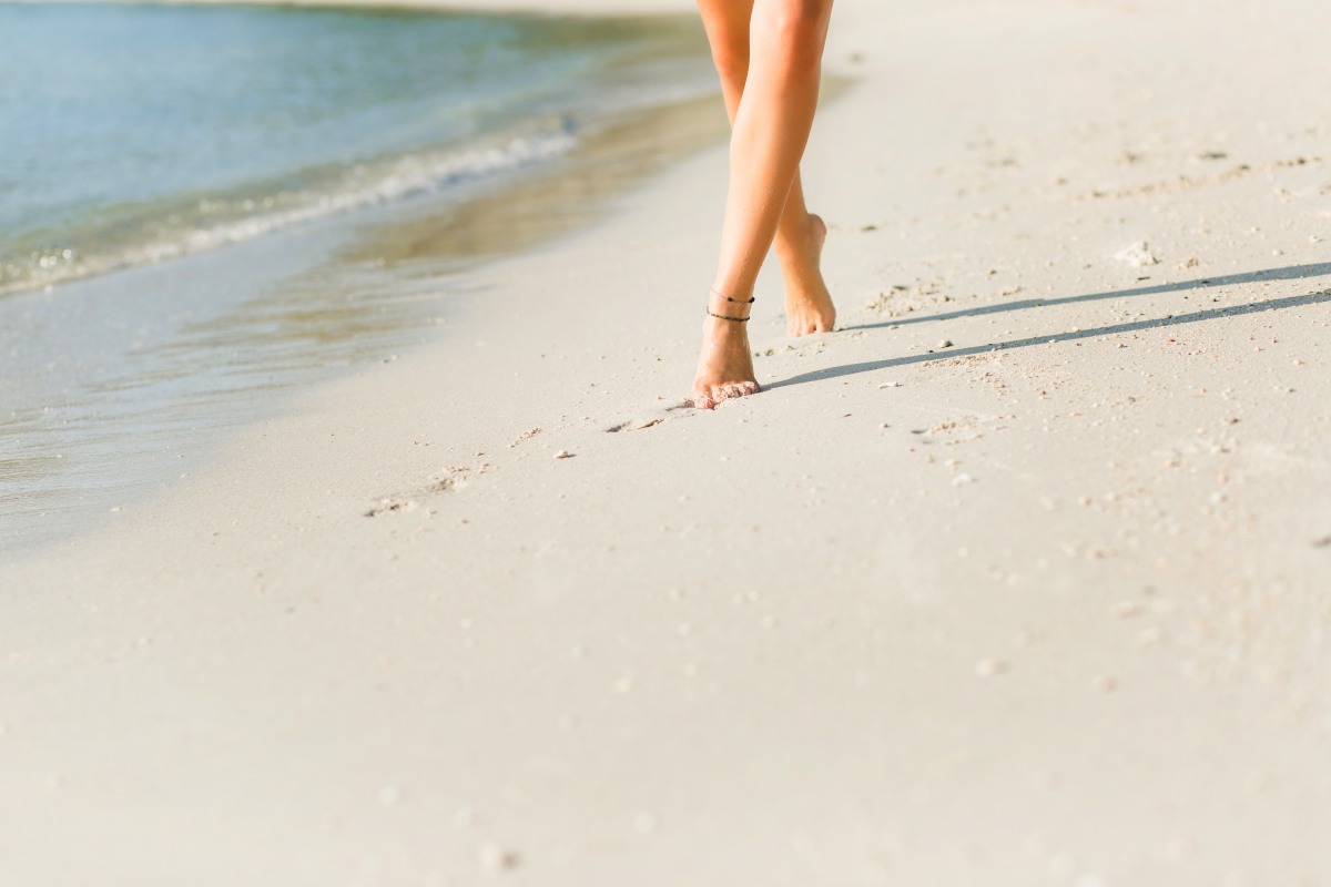 Close-up of tanned slim girl's feet in the sand. She walks near the water. Sand is gold.